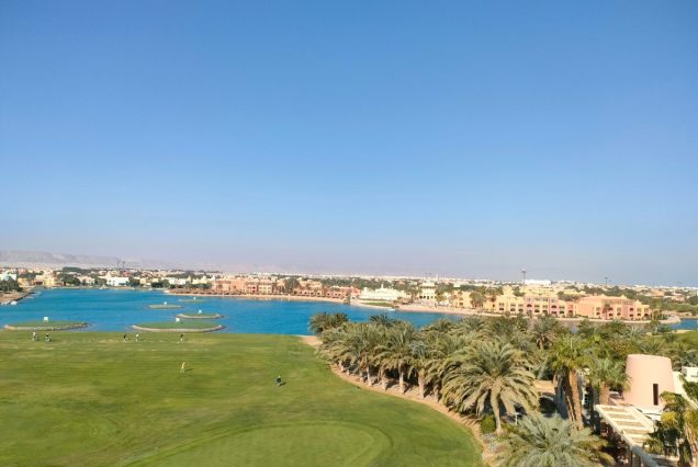 Blick vom Aussichtsturm in El Gouna über einen grünen Golfplatz mit Palmen, eine blaue Lagune mit kleinen Inselgrüns und die Stadt, dahinter Berge